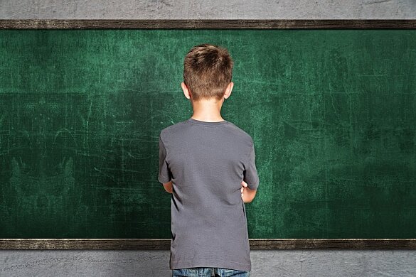 Junge steht vor einer Schultafel mit dem Rücken zur Klasse und verschränkten Armen