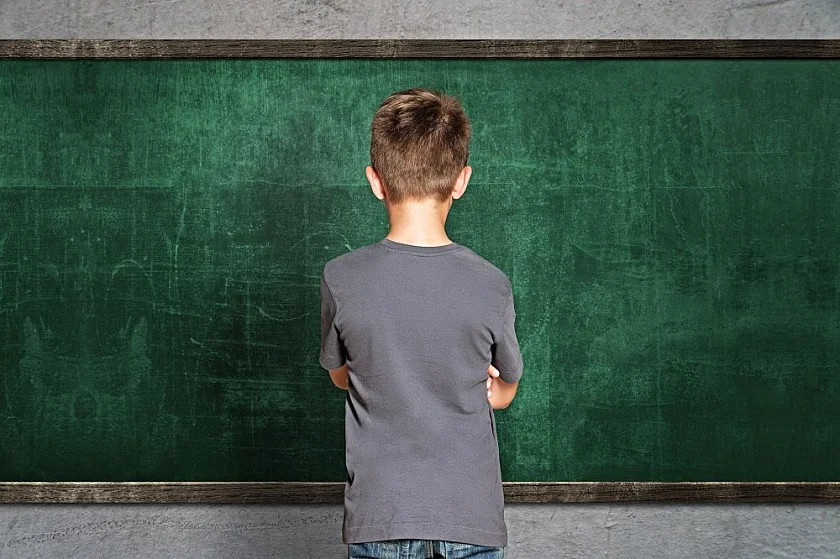 Junge steht vor einer Schultafel mit dem Rücken zur Klasse und verschränkten Armen